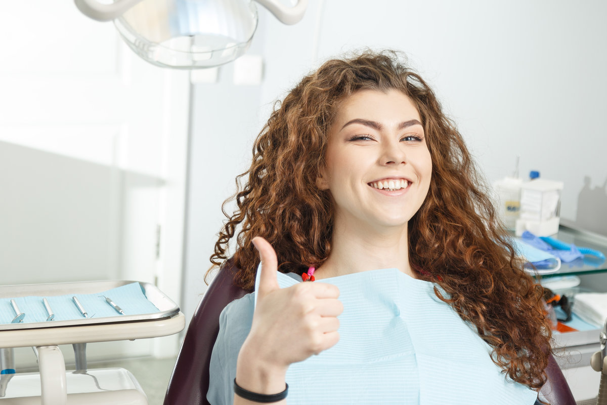 Young female patient visiting dentist office.Beautiful smiling woman with healthy straight white teeth sitting at dental chair and making thumbs up.Dental clinic.Stomatology