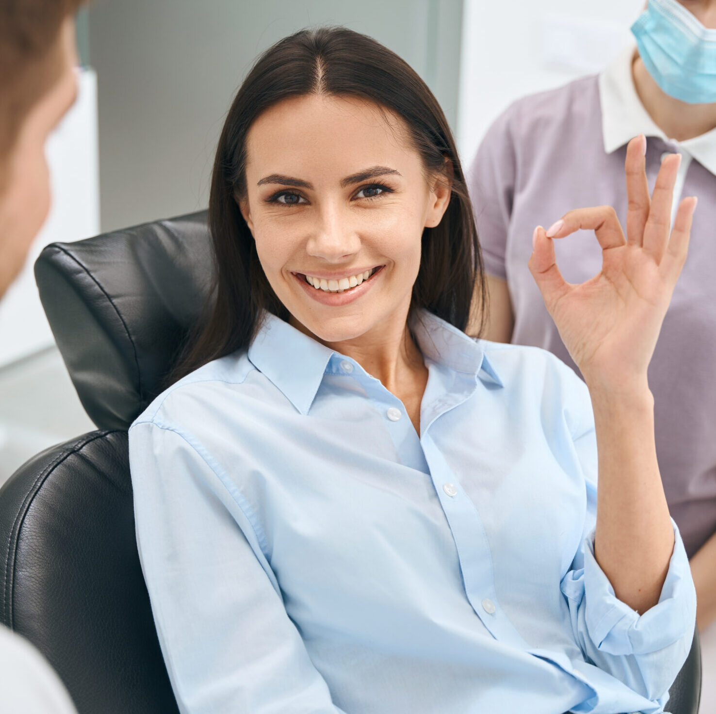 Smiling beautiful woman showing ok gesture sign sitting in orthodontic chair after painless and quality dental treatment, qualified dentist and nurse standing nearby